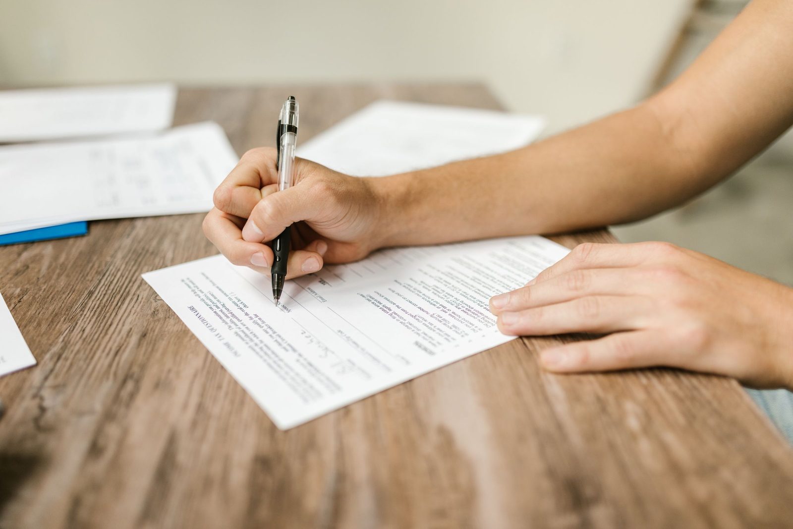 A hand holding a pen, filling out a printed form on a wooden desk